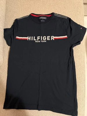 Svart Hilfiger t-shirt med tryck - Snygg svart t-shirt från Tommy Hilfiger med klassiskt tryck 'HILFIGER NEW YORK' framtill och röd-vit rand över bröstet. T-shirten har korta ärmar, rund halsringning och är gjord i mjuk bomull. Perfekt för en clean och stilren look.