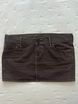 Brun minikjol från Abercrombie - En brun minikjol i jeansmaterial från Abercrombie, dubbla knappar framtill, kontrstsömmar och snygga bakfickor med med små slitningsdetaljer. Perfekt inför sommaren 