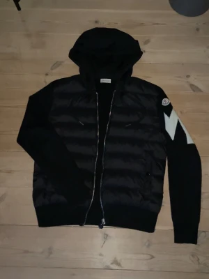 Svart Moncler cardigan med huva - Svart cardiganjacka från Moncler med vadderad framsida, stickade ärmar och huva. Dragkedja framtill och vit detalj på vänster ärm samt Moncler-logga. Perfekt för lager-på-lager och streetwear-stil.