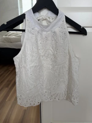 Vit broderad ärmlös linnetopp - Säljer en superfin vit ärmlös linnetopp med broderade detaljer och knyt i nacken. Toppen har ett vackert hålmönster och hög hals, perfekt för varma dagar. Snygg att styla med jeans eller kjol!