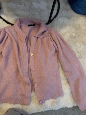 Ljusrosa kofta med krage - Söt ljusrosa kofta med krage och fyra matchande knappar framtill. Koftan har långa ärmar med lätt puff vid axlarna och ribbade muddar. 