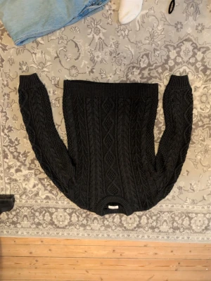 Svart kabelstickad tröja - Säljer en svart kabelstickad tröja med klassiskt flätmönster och rund hals. Tröjan har långa ärmar och breda ribbade muddar vid ärmslut och nederkant. Perfekt för dig som gillar en tidlös och mysig stil.