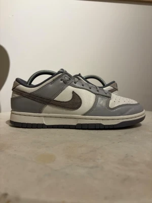 Nike Dunk Low grå/vit sneakers - Nike Dunk Low sneakers i grått och vitt läder med klassisk Swoosh på sidan. Skorna har perforerad tåbox för ventilation, grå snörning och en vit mellansula med grå yttersula. Perfekt för dig som gillar stilrena och tidlösa sneakers. Storlek 41. Skorna är i väldigt fint skick och använda ett fåtal gånger. Skriv till mig om ni har några fler funderingar!