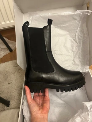 Svarta chunky boots i skinn - Säljer ett par svarta boots med chunky sula och högt skaft. Strl 36, helt nya. Bootsens ovandel är i slätt skinn och har resår på sidorna för enkel påtagning. Skorna köptes för ca 800