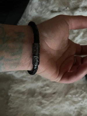 Svart flätat läderarmband med metall - Snyggt svart armband i flätat läder med detaljer i silverfärgad metall. Armbandet har ett robust spänne med ingraverat mönster och passar perfekt för dig som gillar en stilren men edgy look. Köpt på swaroviski och är en del av Laila bagges smycke serie för rock of Swedbank 