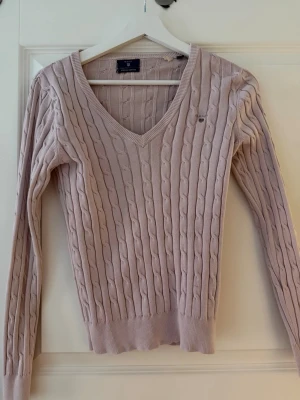 Ljusrosa kabelstickad tröja Gant - Supersnygg ljusrosa kabelstickad tröja från Gant med V-ringning och diskret logga på bröstet. Tröjan har långa ärmar och ribbade muddar vid ärmslut och nederkant. Perfekt för dig som gillar klassisk och stilren look.