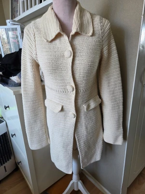 Odd molly kofta - Vacker virkad kofta en elegant krämvit nyans.nkoftan har en klassisk krage och knäppning framtill med virkade knappar. Detaljerade fickor framtill och en dekorativ virkad applikation baktill ger jackan en unik touch. Ärmarna är långa och raka. En siffra '20' är diskret virkad på ryggen, vilket bidrar till dess charm. Strl 0 vet ej om det är xs eller s men sittrr mer som s
