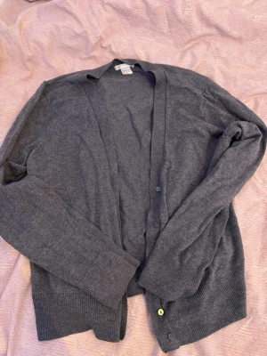 Mörkgrå cardigan från H&M - Mjuk och tunn mörkgrå cardigan från H&M med långa ärmar och ribbade muddar. Klassisk V-ringad modell med knäppning framtill och diskreta knappar. Perfekt att slänga över en t-shirt eller skjorta för en chill look.
