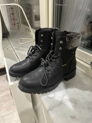 Svarta kängor med foder  - Snygga svarta kängor med grov sula och snörning. Kängorna har ett mjukt grått foder upptill och detaljer i metall. Perfekta för kalla dagar och ger en cool look till din outfit.