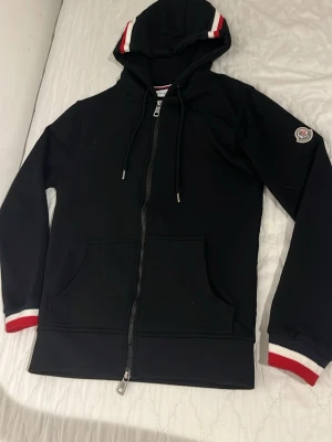 Svart Moncler hoodie med dragkedja - Snygg svart hoodie från Moncler med dragkedja framtill, huva med dragsko och fickor på magen. Detaljer i vitt och rött på muddar och huva samt Moncler-logga på ärmen. Perfekt för en stilren och sportig look.
