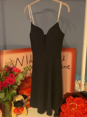 Svart kort klänning med pärlband - Stilren svart kort klänning med djup v-ringning och pärlprydda axelband. Klänningen har en figurnära överdel och lätt utställd kjol, perfekt för en trendig look. Materialet känns mjukt och följsamt mot kroppen. Aldrig använd lappen kvar ❤️