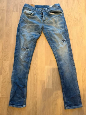Blå slitna jeans med knappar - Ett par blå jeans med slitningar och hål på vänster lår, klassisk femficksmodell och knappgylf. Jeansen har en tvättad look och raka ben. Perfekta för en avslappnad och trendig stil.