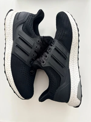 Svarta Adidas Ultra Boost sneakers - Snygga svarta Adidas Ultra Boost sneakers med stickad ovandel i mesh, vita Boost-sulor och svarta plastdetaljer på sidorna. Klassisk tre-randig design och bekväm passform. Perfekta för dig som gillar sportig stil och streetwear.