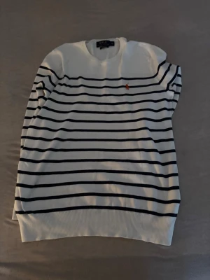 Vit och marinblå randig tröja Polo Ralph Lauren - Klassisk vit tröja från Polo Ralph Lauren med marinblå horisontella ränder och broderad logga i orange på bröstet. Rund halsringning och ribbade muddar vid ärmslut och nederkant. Perfekt för en stilren och avslappnad look passar 160-167 cm