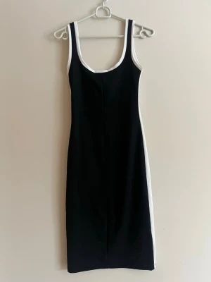 Svart-vit ärmlös klänning - Säljer en svart ärmlös klänning med vita kanter längs sidorna och runt halsringningen. Klänningen har en kroppsnära passform och är i midilängd. Perfekt för dig som gillar en stilren och sportig look.