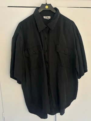 Svart oversized skjorta i silke  - Säljer en svart oversized skjorta från Silk road  med korta ärmar och två bröstfickor med lock och knapp. Skjortan har klassisk krage och knäppning framtill. Perfekt för en avslappnad och cool stil. 100 % silk. #vintage 
