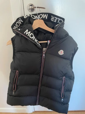 Moncler väst - Snygg svart dunväst från Moncler med stor huva och logotyp på bröstet. Västen har dragkedja framtill, två fickor med dragkedja och Moncler-tryck på huvkanten. Super bra skick bara andvänt några tillfällen. Storlek 170-176 typ till en 15-16 åring.