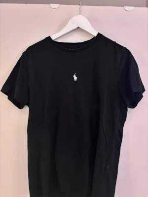 Svart t-shirt från Polo Ralph Lauren - Klassisk svart t-shirt från Polo Ralph Lauren med den ikoniska vita logon broderad på bröstet. T-shirten har rund halsringning och korta ärmar. Tillverkad i mjuk bomull för en skön känsla och enkel stil.