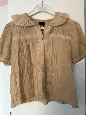 Beige blus med volangkrage och brodyr - Oanvänd, söt, beige blus med korta puffärmar, volangkrage och knappar framtill. Blusen har broderade detaljer och ett lätt skrynklat tyg som ger en avslappnad känsla. Perfekt för dig som gillar romantisk och vintageinspirerad stil.
