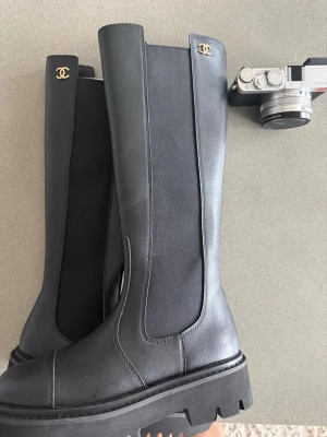 Svarta höga stövlar från C - Säljer ett par svarta höga stövlar från C med chunky sula och guldfärgade logodetaljer upptill. Stövlarna är i skinn med elastiska partier på sidorna för skön passform. Perfekta för dig som vill ha en lyxig och trendig look.