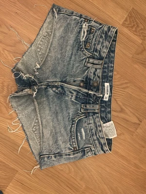 Blå denimshorts från Pull&Bear - Säljer ett par blå denimshorts från Pull&Bear med råa kanter och slitna detaljer. Shortsen har låg midja, dubbla knappar och klassiska fem fickor. Perfekta för sommaren och ger en avslappnad vibe. 