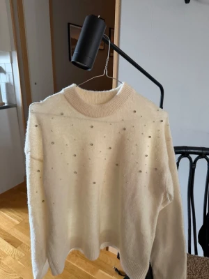 Vit stickad tröja med pärldetaljer - Säljer en krämvit stickad tröja med rund hals och långa ärmar. Tröjan har små pärlor och stenar som dekoration framtill och upptill på ryggen. Mjuk och fluffig känsla, perfekt för dig som gillar en enkel men ändå lite extra look.