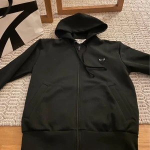 CDG Zip Hoodie Svart - Säljer en stilren svart hoodie från Comme des Garçons Play med en ikonisk hjärtdetalj på bröstet. Tröjan har dragkedja och en stor hjärtdetalj på ryggen. Perfekt för en casual look. Storlek S och i fint skick. 