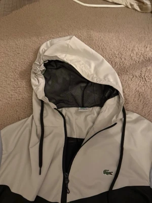 Lacoste vindjacka med huva - Snygg vindjacka från Lacoste i svart, vitt och ljusgrått. Jackan har huva med dragsko, meshfoder och dragkedja framtill. Stora Lacoste-loggor på båda ärmarna och den klassiska krokodilen på bröstet. Perfekt för en sportig och trendig look.