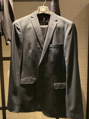 Mörkblå kavaj från Tiger of Sweden - Stilren mörkblå kavaj från Tiger of Sweden med klassisk passform, två framfickor med lock och en bröstficka. Kavajen har smala slag och är tillverkad i ett slätt tyg som ger en elegant look. Perfekt för dig som vill ha en tidlös och snygg outfit. Sen kommer även byxor och väst med