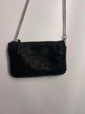 Svart kuvertväska med kedja - Snygg svart kuvertväska med glansig yta och silverfärgade nitar längs kanterna. Väskan har en lång silverkedja som axelrem och stängs med dragkedja upptill. Perfekt för dig som gillar en edgy och stilren look.