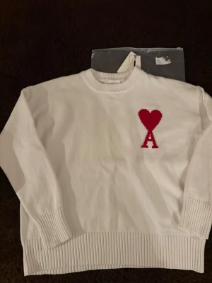 Vit stickad tröja från Ami Paris - Vit stickad tröja från Ami Paris med rund halsringning och ribbade muddar. På bröstet finns ett rött hjärta och bokstaven 'A' i intarsia. Tröjan har en avslappnad passform och är tillverkad i mjukt material, perfekt för en stilren look.