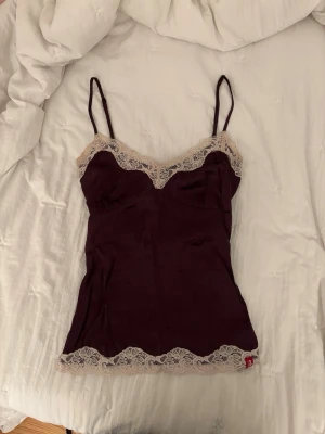 Mörklila linnetopp med spets - Supersöt mörklila linnetopp med tunna axelband och beige spetsdetaljer längs urringning och nederkant. Sitter figurnära och har en feminin, romantisk vibe. Perfekt att styla med jeans eller kjol för en trendig look.