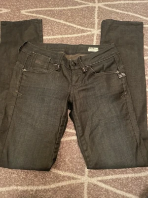 Mörkgrå jeans från G-Star Raw - Snygga mörkgrå jeans från G-Star Raw med raka ben och klassisk femficksmodell. Jeansen har detaljerade sömmar och en läderpatch med logga bak i midjan. Tillverkade i ett slitstarkt denimtyg som ger en cool och tidlös look. Liten skada, se sista bilden