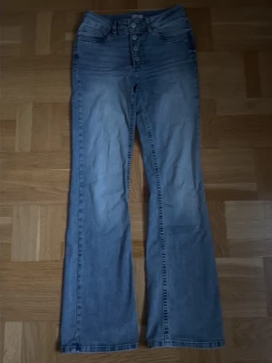 Ljusblå bootcut jeans med knappar - Säljer ett par ljusblå bootcut jeans med medium midja och knappgylf. Jeansen har klassiska fem fickor och är tillverkade i denim. Perfekta för dig som gillar retrostil och vill ha ett par bekväma jeans med snygg passform.