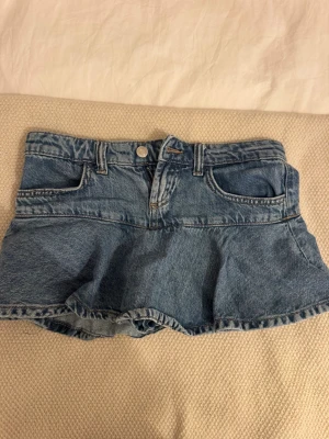 Blå jeansskort från Bershka - Supersnygg blå jeanskjol från Bershka i klassisk denim med fem fickor och knappgylf. Kjolen har en utställd kjoldel över shortsen och är gjord i ett mjukt bomullsdenim. 