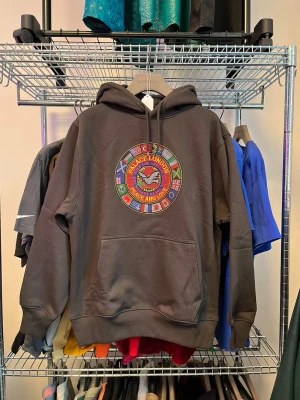 Mörkgrå hoodie från Palace - Säljer en mörkgrå hoodie från Palace med ett stort, färgglatt broderat emblem på bröstet med texten 'Peace and Love' och flera flaggor runt om. Hoodien har huva med dragsko och en stor magficka. Perfekt för dig som gillar streetwear och vill sticka ut.