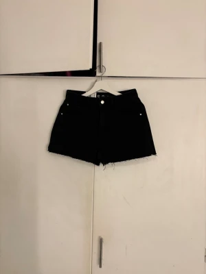 Svarta denimshorts från Gina Tricot - Svarta jeansshorts från Gina Tricot med rå kant nertill och klassisk femficksmodell. Shortsen har hög midja och är tillverkade i bomull för en skön och avslappnad look. Perfekta till sommaren och lätta att matcha med olika stilar.