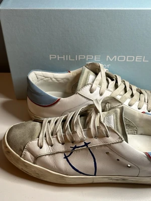 Philippe Model vita sneakers - PRIS KAN DISKUTERAS! Snygga vita Philippe Model sneakers med grå mockadetaljer på tån, blå och röda detaljer vid hälen samt blå logga på sidan. Skorna har vita snören och en slitstark gummisula. Perfekta för dig som gillar en avslappnad och trendig stil!
