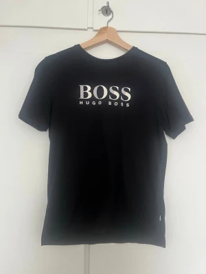 Svart Hugo Boss t-shirt med logga - Snygg svart t-shirt från Hugo Boss med stor vit logga på bröstet. Klassisk rund halsringning och korta ärmar. Tillverkad i mjuk bomull som känns skön mot huden. Perfekt för en clean och stilren look.