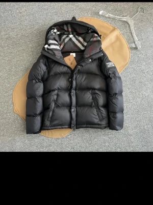 Svart pufferjacka från Burberry - Snygg svart jacka från Burberry med avtagbara ärmar och huva. Västen har klassiskt rutigt foder i huvan, två framfickor med dragkedja och tryckknappar framtill. Märkespatch på ena ärmen och cool, puffig design som håller dig varm.