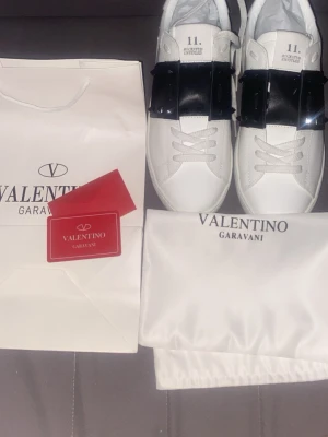 Valentino Garavani vita sneakers - Säljer ett par helt nya Valentino Garavani Open sneakers i vitt skinn med bred svart rem och nitar på sidorna. Skorna kommer med dust bag och allt annat!.