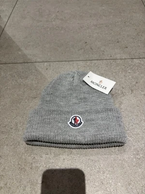 Grå Moncler mössa i ribbstickad ull - Snygg grå ribbstickad mössa från Moncler med klassisk uppvikt kant och broderad logotyp framtill. Perfekt accessoar för att hålla värmen och samtidigt se trendig ut. Materialet är mjuk ull som känns skönt mot huden.
