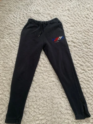 Svarta mjukisbyxor från Trapstar - Säljer ett par svarta mjukisbyxor från Trapstar med broderad logga i blått, rött och vitt på vänster ben. Byxorna har dragsko i midjan, muddar vid bensluten och dragkedja vid anklarna. Perfekta för en chill och streetwear-inspirerad look.