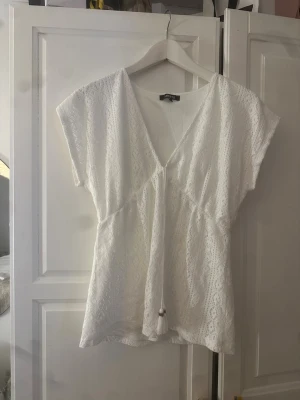 Vit spetsblus med knyt från Liberto - Söt vit blus från Liberto i spets med v-ringning och knyt framtill. Blusen har korta ärmar och ett luftigt, avslappnat snitt som passar perfekt till sommaren. Detaljerad spets ger ett romantiskt intryck och den är lätt att matcha med olika stilar.