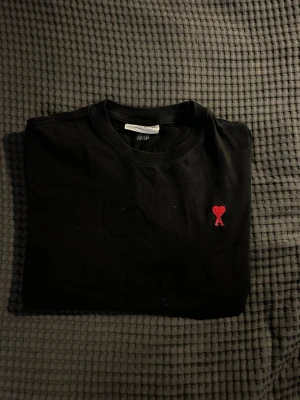 Svart t-shirt från Ami Paris - Svart t-shirt från Ami Paris med klassisk passform och korta ärmar. På bröstet finns ett litet rött broderat hjärta med bokstaven A. Tillverkad i mjuk bomull och perfekt för en clean och stilren look.