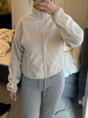 Krämvit fleecejacka med dragkedja - Mysig beige fleecejacka med hög krage och hel dragkedja framtill. Jackan har två sidofickor med dragkedja och en avslappnad passform.  