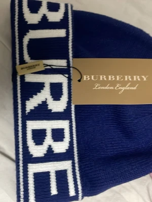 Burberry mössa med logotyp - Två snygga mössor från Burberry, en i blått med vit logotyp och en i grått med svart logotyp. Klassisk ribbstickad design med bred uppvikt kant och tydlig BURBERRY-text framtill. Perfekt accessoar för att lyfta din streetstyle.