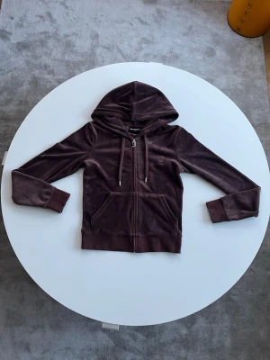 Bruna velour hoodie från JUICY COUTURE - Mysig brun hoodie från Juicy Couture  med dragkedja och huva💕. Klassisk broderad JC-logga på bröstet och snygg J-dragkedja. Fickor framtill och långärmad modell. Den är andvänd men i fint skick🥰 