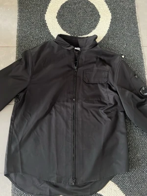 Svart overshirt från C.P. Company - Snygg svart overshirt från C.P. Company med dragkedja framtill, bröstficka och ikonisk linsdetalj på ärmen. Overshirten har en modern, rak passform och är tillverkad i ett slätt, tekniskt material som ger en clean look.