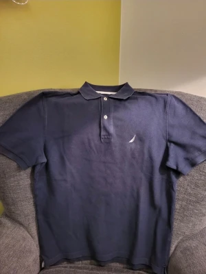 Marinblå pikétröja från Nautica S - Klassisk marinblå pikétröja från Nautica med vit broderad logga på bröstet. Tröjan har korta ärmar, krage och två knappar framtill. Tillverkad i bomull för en bekväm känsla och stilren look.